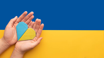 Stand with Ukraine concept. Hands holding blue and yellow heart on Ukraine flag background. Pray for Ukraine peace.