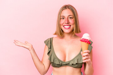 Young caucasian woman wearing a bikini and holding an ice cream isolated on pink background showing a copy space on a palm and holding another hand on waist.