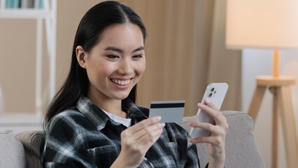 Smiling asian ethnic woman customer user holding bank credit card with virtual money typing smartphone at home. Happy multiethnic female shopper using easy mobile payments app making purchase online