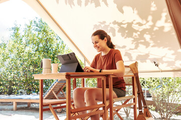 Woman using a laptop on a cozy glamping tent in a sunny day. Luxury camping tent for outdoor summer...
