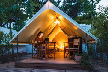 Cozy open glamping tent with light inside and a woman using a laptop during dusk. Luxury camping tent for outdoor summer lifestyle concept