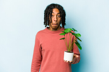 Young African American man holding a plant isolated on blue background shrugs shoulders and open...
