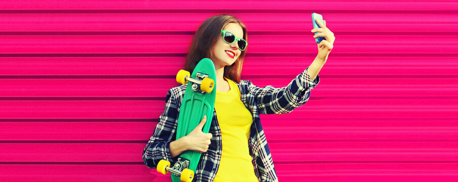 Summer portrait of happy smiling young woman model taking selfie by smartphone with skateboard on colorful pink background