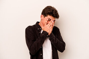 Young caucasian man isolated on white background blink at the camera through fingers, embarrassed covering face.