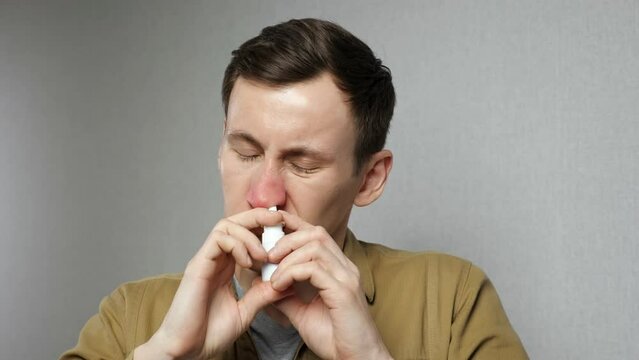 Allergic man sprays nasal medication into nostrils to get relief standing on grey background. Male person suffers from pollen allergy close view