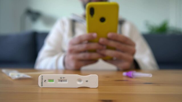 Man using his smart phone after make antigen diagnostic test with a negative result of Covid-19 at home. Person is typing smartphone on wood table with kit for self testing to check Coronavirus