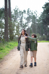Satisfied mother and son walking in forest. Dark-haired wo and boy in coats walking on cloudy day, pointing. Family, nature, leisure concept