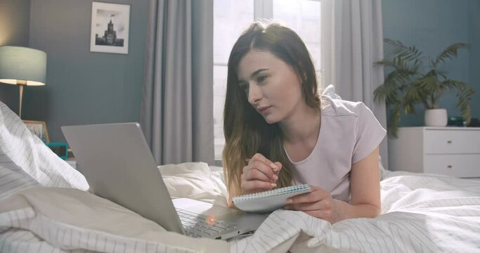 Attractive caucasian female lying on the bed and using laptop notebook to work. Pretty female styding, making notes in paper notebook in bedroom.