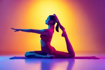  Portrait of young sportive woman in black tracksuit doing yoga and stretching exercises isolated over gradient pink and yellow background in neon © Lustre