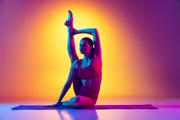 Portrait of young sportive girl doing yoga exercises, sitting, rising leg upwards isolated over gradient pink and yellow background in neon.