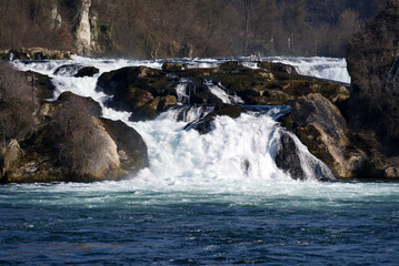Famous Rhine Falls with rocks and splashing water on a sunny spring day, focus on background. Photo taken March 7th, 2022, Zurich, Switzerland.