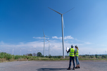 Engineer wearing uniform ,helmet hold document inspection work in wind turbine farms rotation to generate electricity energy. Green ecological power energy generation wind sustainable energy concept.