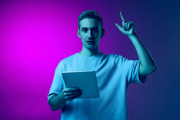 Portrait of young man with tablet rising finger up symbolizing new ideas isolated over gradient pink and purple background in blue neon