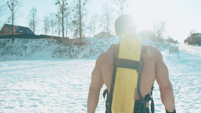 A muscular man with a naked torso and ballon compressed air drager on his back goes through the snow in the park. Back view