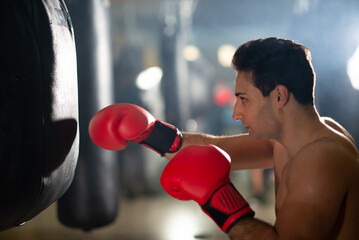 Boxer doing punch training in gym. Young shirtless man in boxing gloves kicking punching bag against light. Sport concept