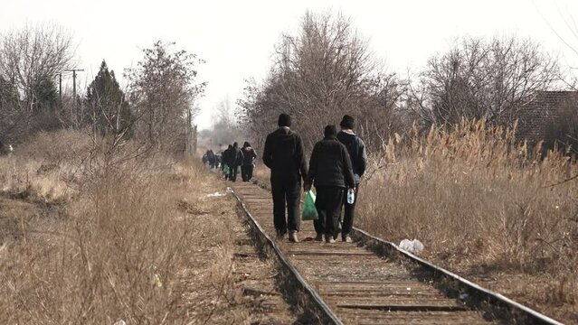 A telephoto video clip of dozens refugees walking on a train track in the distance, carrying all their belongings with them, in a cold, sunny winter day. some heat haze is apparent.