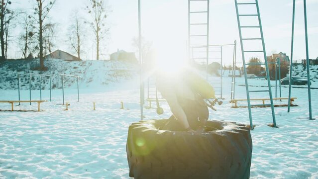 A muscular man with a naked torso with a drager on his back does strength training in a winter park and carries a large tire in his hands