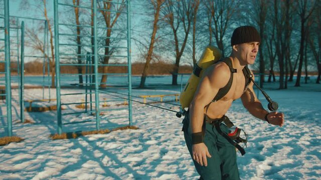 Young muscular man training in a winter park and doing a resistance exercise with a powerlifting band and with a compressed air bottle Drager