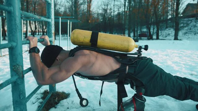 Muscular man without a t-shirt with a ballon compressed air drager on his back is training in a winter park and doing push-ups from the crossbar