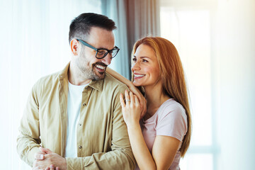 Smiling couple hugging each other and standing near window while looking outside. Happy and romantic man embracing hispanic wife from behind while standing at home with copy space. 