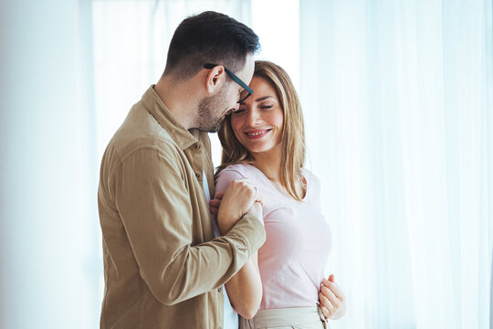 Smiling couple hugging each other and standing near window while looking outside. Happy and romantic man embracing hispanic wife from behind while standing at home with copy space. 