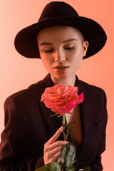 Fototapeta premium charming woman in black brim hat holding pink rose on coral background.