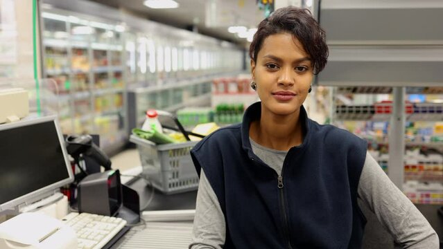 Portrait of young female cashier posing and smiling at supermarket checkout spbi. 4k Close view of beautiful african american woman looks forward and poses with smile, has work break and sits at desk