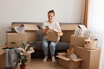 Image of smiling attractive woman wearing white t shirt and jeans sitting on sofa during relocating to a new house and using smart phone, holding carton parcel.