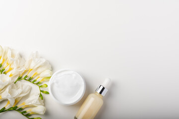 Obraz na płótnie Canvas White cosmetic serum with a pipette in a glass bottle and cream jar, bouquet of white freesias on a white background.