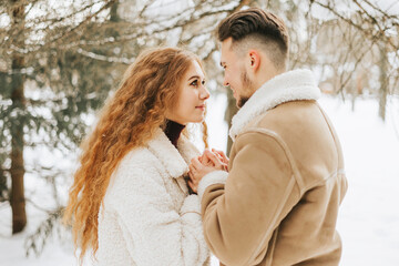 Fototapeta premium young couple in love man and woman in fashion stylish winter clothes standing in snowy pine forest and having fun spending time together, concept of valentine's day and newlyweds, tenderness and love