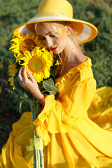 happy girl in a yellow dress holds sunflower flowers in a field of sunflowers against the sky....