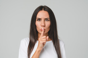 Top secret. Anonymous information. Caucasian young serious woman showing silent gesture, finger on lips, making hush isolated in grey background