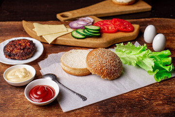 Cooking hamburger or cheeseburger. Different ingredients for a classic hamburger. Grilled meat, vegetables, greens, sauces near a sesame bun. Wooden background. Hamburger day.
