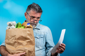 Surprised mature man looking at store receipt after shopping, holding a paper bag with healthy...