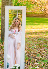 Side view of stunning and pensive girl with beautiful hairdo and makeup, dressed in elegance pink gown which adorned by flowers, standing in front of the mirror