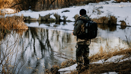 A fisherman with a fishing rod catches fish on the bank of a snow-covered river in early spring