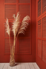 high pampas grass in a vase against a terracotta-red wooden cabinet with stacked doors like blinds....