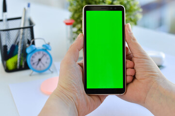 Young woman at home uses green mock-up screen smartphone. She's sitting at a table with a clock and...