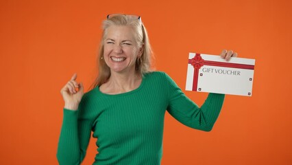 Happy mature blonde woman 40s 50s dancing with gift certificate voucher thumbs up winner gesture isolated on orange color background studio portrait.