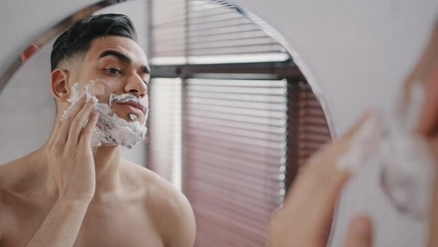 Hispanic male bearded arabian arab indian millennial 30s man model using white foam shaving gel natural cosmetics smears beard cheeks bathroom looking at mirror. Guy prepare for shaves beard mustache