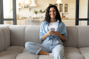 Happy young woman sitting on couch in living room and using cellphone, checking new mobile application at home