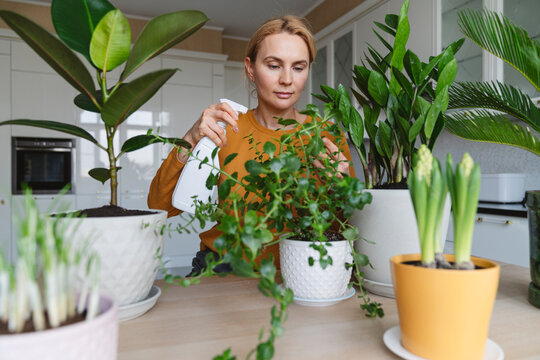 A nice woman in a yellow jacket is watering her home plants.