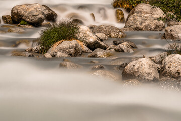 Long exposure and close-up detail of a river flowing between stones and clumps of grass. 