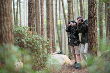 Glad mother and son spending time in forest. Woman and son in casual clothes with cameras peeking from behind trees. Hobby, family, nature, photography concept