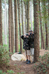 Happy mother and son spending time in forest. Woman and son in casual clothes with cameras peeking from behind trees. Hobby, family, nature, photography concept