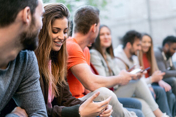 Group of trendy young people chatting together sitting on a bench outdoors. Students having fun together. Focus on the blonde girl