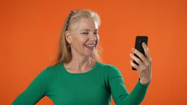 Portrait of smiling mature woman posing holding phone video chat isolated on orange background.