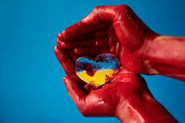 woman's hands on a blue background holding a paper cutout of a heart painted in the colors of the...