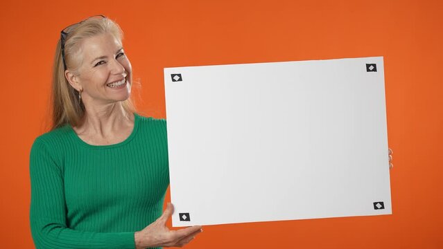 Excited smiling mature woman in green shirt isolated on orange background holding white blank sign board with place for text.