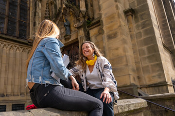 Two young Caucasian female friends in the city together with a book sitting next to a church, enjoying the spring weekend